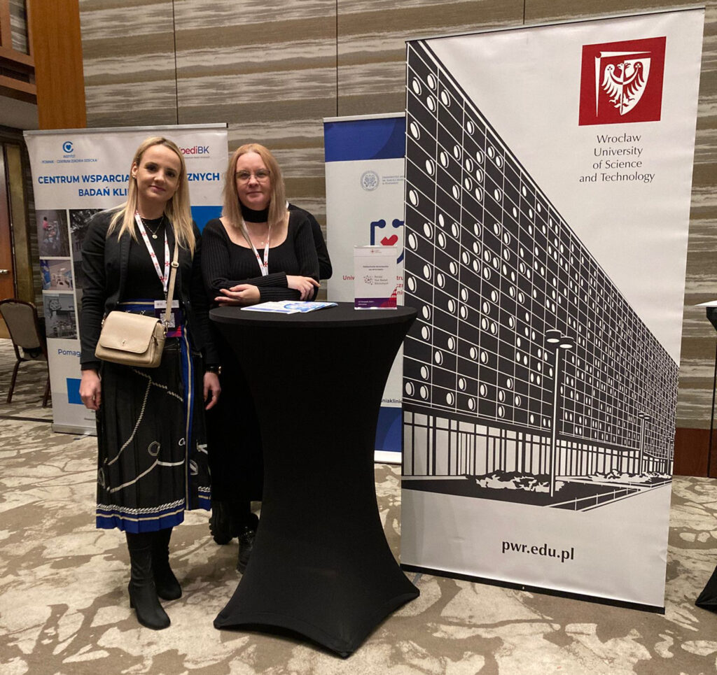 Dwie kobiety przy stoliku informacyjnym obok roll-upów uczelni.