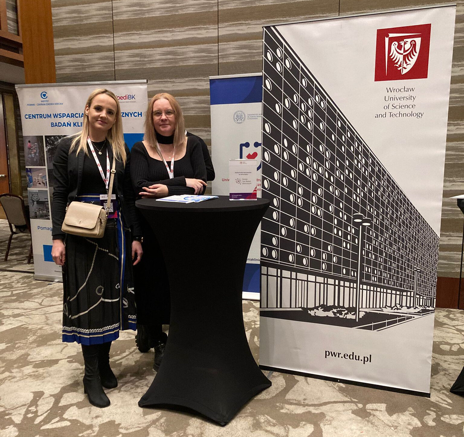 Dwie kobiety przy stoliku informacyjnym obok roll-upów uczelni.