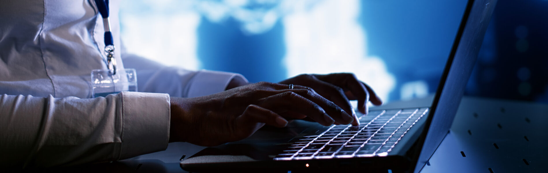 Osoba pisząca na laptopie w ciemnym pomieszczeniu z niebieskim tłem technologii.